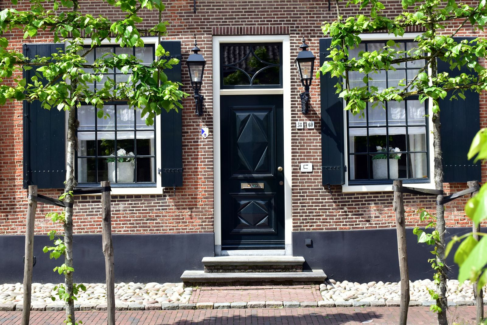 Bakstenen gevel met een zwarte voordeur tussen twee ramen met luiken en lantaarns